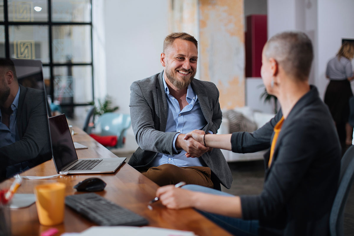 A business professional and happy client shake hands while seated at a long wooden table near a laptop computer.