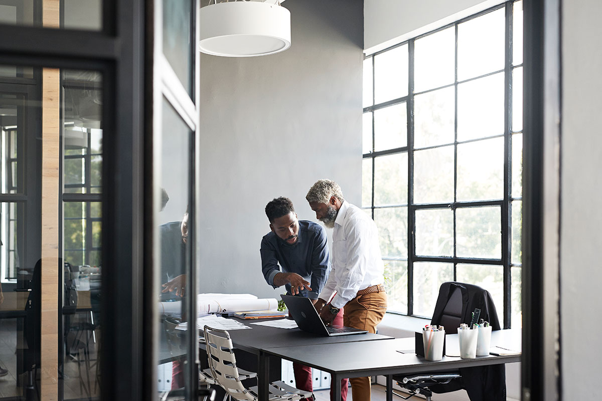 Two casual professionals stand in a bright room, having a discussion over a table containing a laptop and rolled drawings.