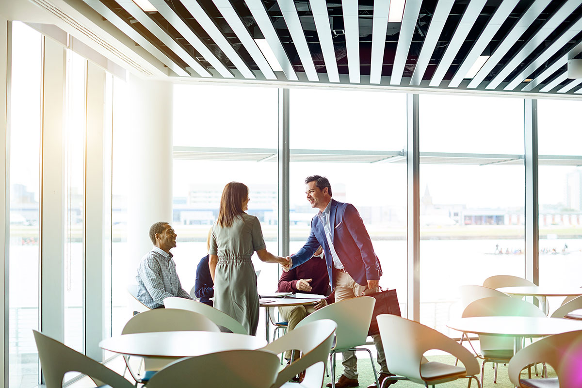 A commercial property insurance professional meets and shakes hands with clients in a bright glass-walled room with several cafe-style tables.
